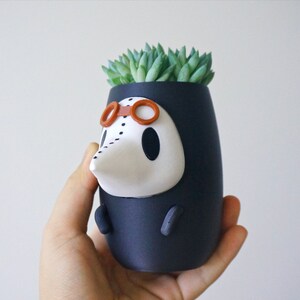 May include: A black and white ceramic planter shaped like a character wearing a black hooded robe, goggles, and a white mask. The planter has a small succulent plant growing out of the top.