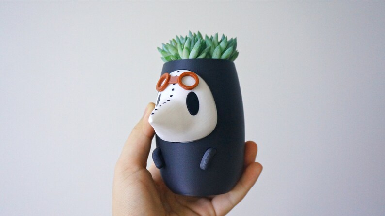 May include: A black and white ceramic planter shaped like a character wearing a black hooded robe, goggles, and a white mask. The planter has a small succulent plant growing out of the top.