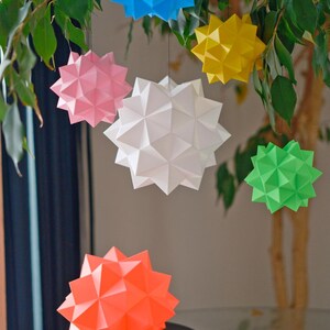 Pode incluir: Uma cole&ccedil;&atilde;o de oito estrelas de papel origami em v&aacute;rias cores, incluindo azul, rosa, amarelo, branco, verde e laranja. As estrelas est&atilde;o penduradas em um galho de &aacute;rvore e algumas est&atilde;o sentadas em uma mesa.