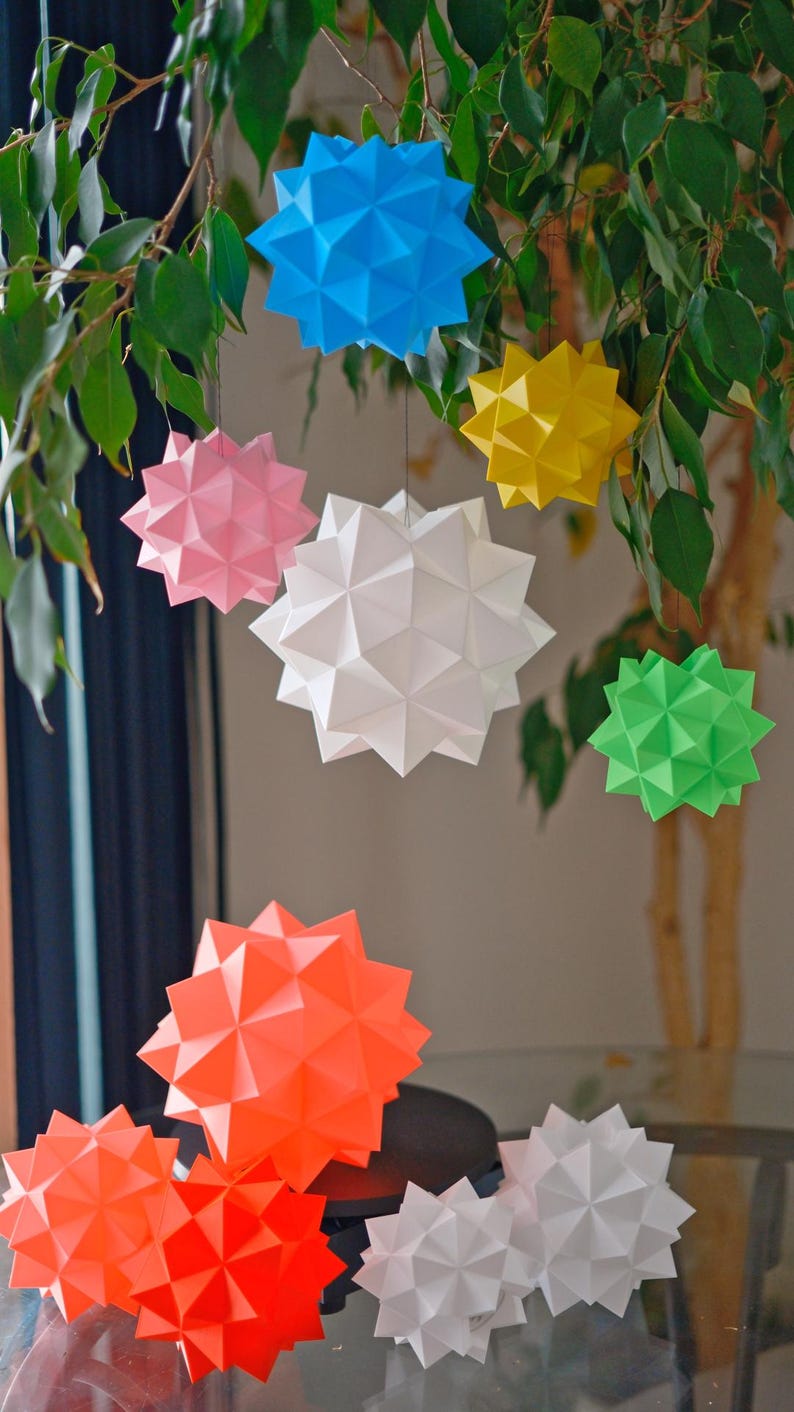 Pode incluir: Uma cole&ccedil;&atilde;o de oito estrelas de papel origami em v&aacute;rias cores, incluindo azul, rosa, amarelo, branco, verde e laranja. As estrelas est&atilde;o penduradas em um galho de &aacute;rvore e algumas est&atilde;o sentadas em uma mesa.