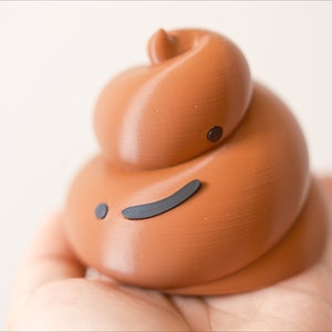 May include: A brown, 3D printed poop emoji with a black, winking face. The poop is being held in a hand.