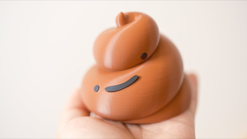 May include: A brown, 3D printed poop emoji with a black, winking face. The poop is being held in a hand.