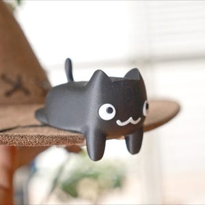 May include: A black cat ornament with white eyes and a smiling mouth sits on a brown hat. The hat has a wide brim and decorative stitching. The cat is a cute, minimalist design.