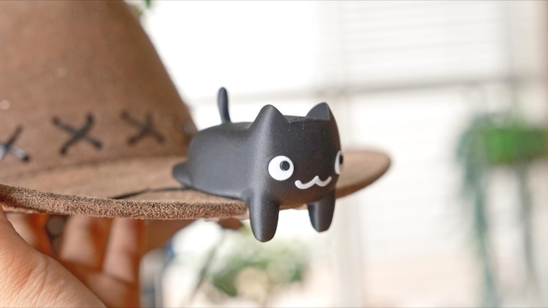 May include: A black cat ornament with white eyes and a smiling mouth sits on a brown hat. The hat has a wide brim and decorative stitching. The cat is a cute, minimalist design.
