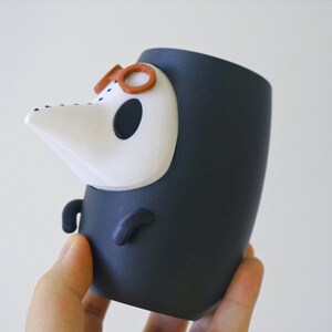 May include: A black and white ceramic figurine of a character wearing a black robe and a white mask with a long nose. The character has brown glasses on top of the mask.