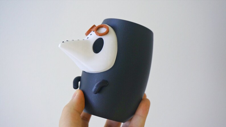 May include: A black and white ceramic figurine of a character wearing a black robe and a white mask with a long nose. The character has brown glasses on top of the mask.