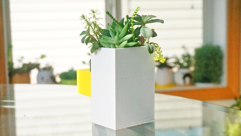 May include: A white planter with a yellow accent holds a variety of green succulents. The planter is sitting on a glass table.