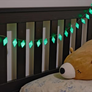 Puede incluir: Una cadena de luces verdes brillantes en forma de diamante cuelga sobre una cabecera de madera oscura. Una gran almohada con forma de oso marrón descansa sobre una almohada estampada, creando una escena acogedora.