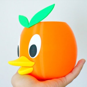 May include: Orange-colored ceramic planter shaped like a cartoon duck with a yellow beak and green leaves on top.