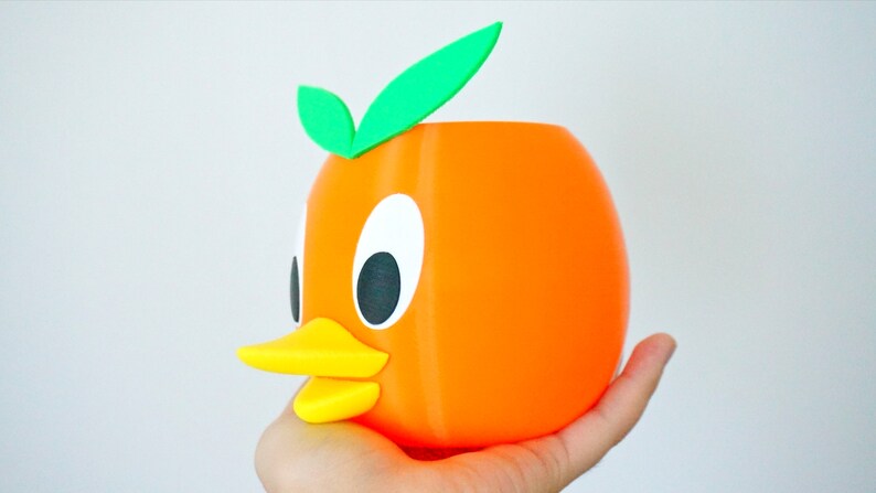 May include: Orange-colored ceramic planter shaped like a cartoon duck with a yellow beak and green leaves on top.