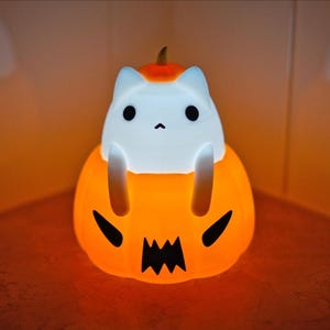 May include: A glowing Halloween-themed night light featuring a white cat figure perched atop an orange pumpkin. The pumpkin has a carved jack-o'-lantern face. The cat has black eyes and a small nose. The light emits a warm, inviting glow.
