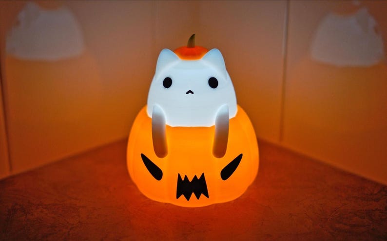 May include: A glowing Halloween-themed night light featuring a white cat figure perched atop an orange pumpkin. The pumpkin has a carved jack-o'-lantern face. The cat has black eyes and a small nose. The light emits a warm, inviting glow.