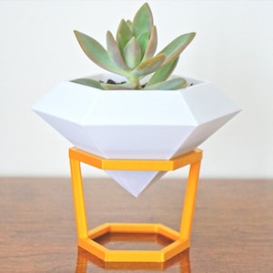 May include: A white diamond-shaped planter with a succulent plant inside, sitting on a yellow hexagonal stand.
