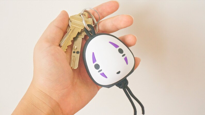 May include: A keychain featuring a white, oval-shaped charm with a black border and purple accents, resembling a character from an animated film. The keychain is attached to a set of keys.