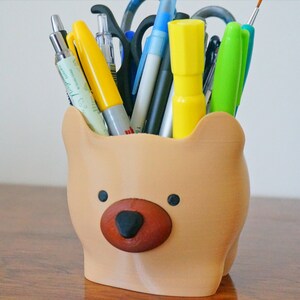Puede incluir: Un organizador de escritorio con forma de oso marr&oacute;n con nariz marr&oacute;n y ojos negros, lleno de varios bol&iacute;grafos, l&aacute;pices y rotuladores.