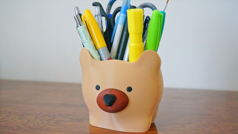 Puede incluir: Un organizador de escritorio con forma de oso marr&oacute;n con nariz marr&oacute;n y ojos negros, lleno de varios bol&iacute;grafos, l&aacute;pices y rotuladores.