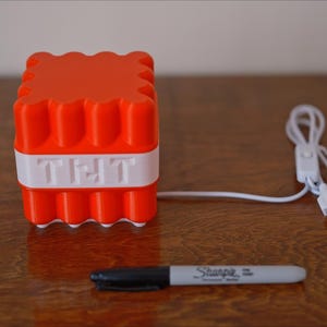 May include: A red and white 3D printed cube with the word "TNT" on the front. The cube is connected to a white USB cable with a switch.