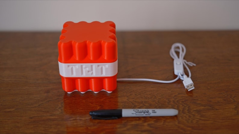 May include: A red and white 3D printed cube with the word "TNT" on the front. The cube is connected to a white USB cable with a switch.