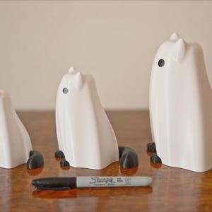 May include: Three white ceramic ghost figurines with black eyes and black feet. The ghosts are different sizes, with the largest ghost being about 6 inches tall.