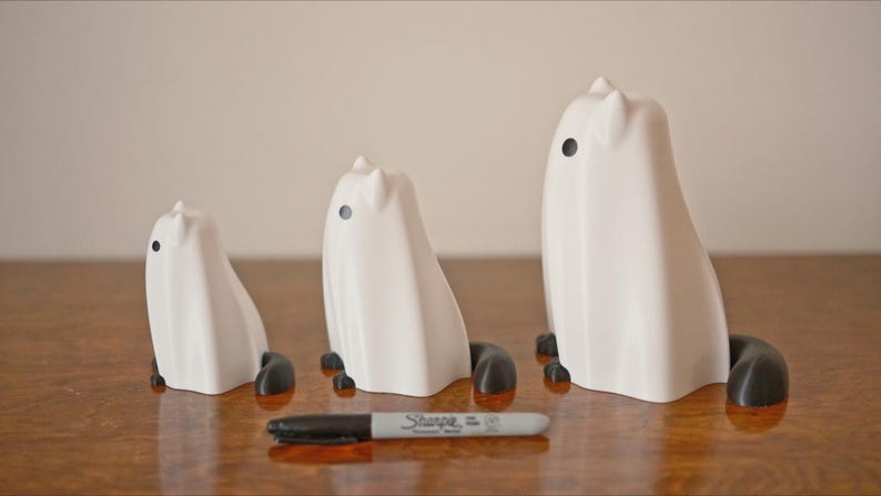 May include: Three white ceramic ghost figurines with black eyes and black feet. The ghosts are different sizes, with the largest ghost being about 6 inches tall.