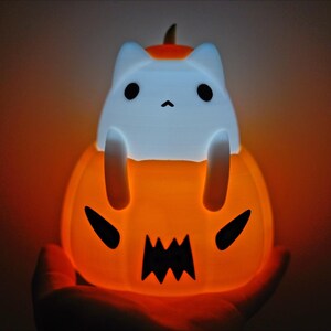 May include: A glowing orange Halloween-themed night light shaped like a jack-o'-lantern with a white cat figure on top. The cat has black eyes and is sitting inside the pumpkin. The pumpkin has a carved face with a jagged mouth.