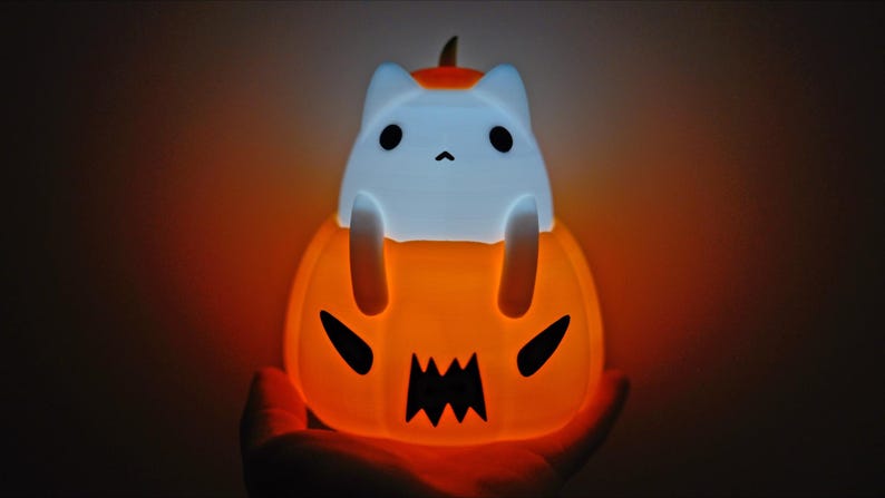 May include: A glowing orange Halloween-themed night light shaped like a jack-o'-lantern with a white cat figure on top. The cat has black eyes and is sitting inside the pumpkin. The pumpkin has a carved face with a jagged mouth.