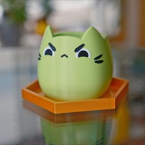 May include: A green cat-shaped planter with a grumpy expression sits on a hexagonal orange coaster.