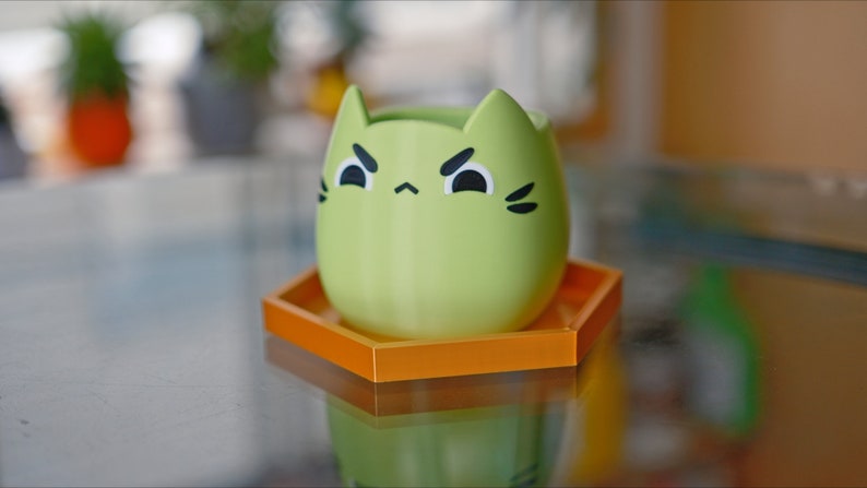 May include: A green cat-shaped planter with a grumpy expression sits on a hexagonal orange coaster.