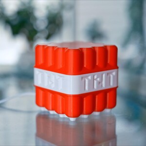 May include: A red and white cube-shaped object with the text "TNT" on the front and "TNT" on the back. The object is made of plastic and has a textured surface.
