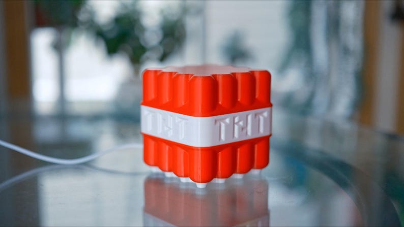 May include: A red and white cube-shaped object with the text "TNT" on the front and "TNT" on the back. The object is made of plastic and has a textured surface.