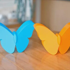May include: Two butterfly-shaped decorations, one light blue and one orange, sit on a wooden surface. The butterflies have a simple, stylized design with rounded wings and a central body. The background is blurred.