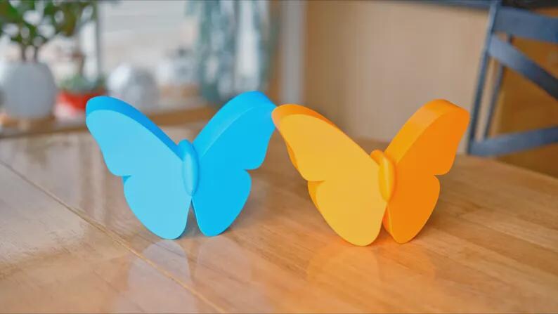 May include: Two butterfly-shaped decorations, one light blue and one orange, sit on a wooden surface. The butterflies have a simple, stylized design with rounded wings and a central body. The background is blurred.