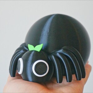 May include: A black and green 3D printed spider toy with large white eyes and a green bow on its head.
