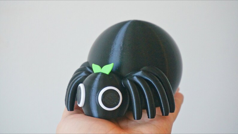 May include: A black and green 3D printed spider toy with large white eyes and a green bow on its head.
