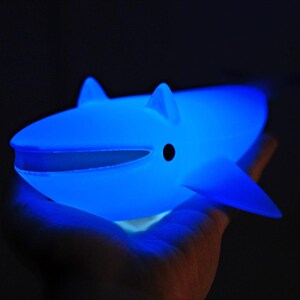 May include: A glowing blue whale-shaped night light. The toy has a smooth surface, fins, and a small black eye. The whale is held in a hand against a dark background, emitting a soft, ambient glow.