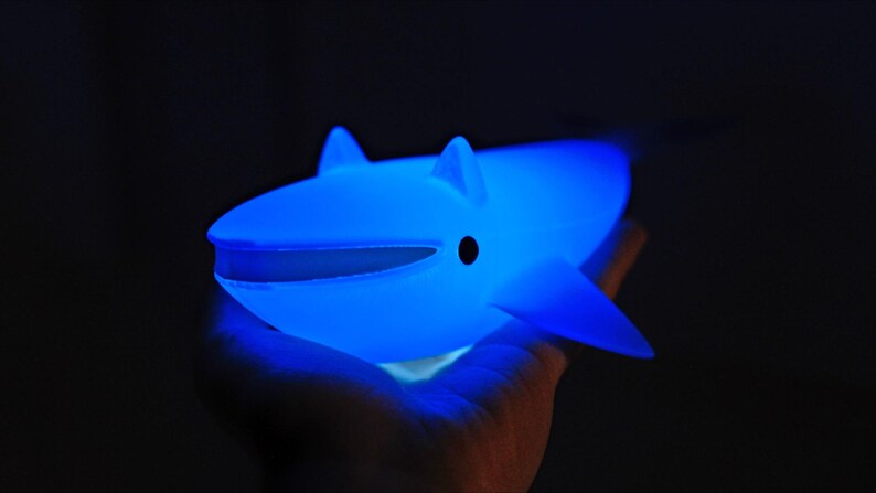 May include: A glowing blue whale-shaped night light. The toy has a smooth surface, fins, and a small black eye. The whale is held in a hand against a dark background, emitting a soft, ambient glow.