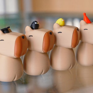May include: Four small, beige, cartoon-like figurines with different hats. The figurines have brown noses and closed eyes. The hats are a black graduation cap, a black bird, a yellow bird, and a red party hat.