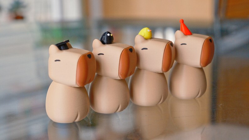 May include: Four small, beige, cartoon-like figurines with different hats. The figurines have brown noses and closed eyes. The hats are a black graduation cap, a black bird, a yellow bird, and a red party hat.