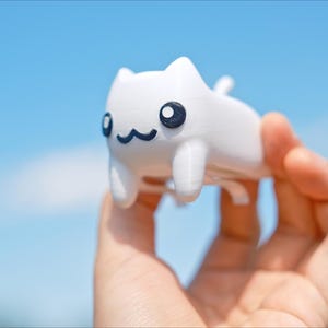 May include: A white cat figurine with black eyes and a smiling mouth is held in a hand against a blue sky. The cat has a simple, cartoonish design, with small ears and a tail. The figurine is a cute and whimsical item.