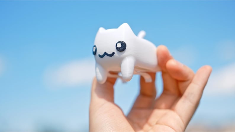 May include: A white cat figurine with black eyes and a smiling mouth is held in a hand against a blue sky. The cat has a simple, cartoonish design, with small ears and a tail. The figurine is a cute and whimsical item.