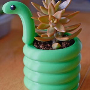 May include: A green snake-shaped planter with a succulent plant. The planter is a vibrant green, coiled to resemble a snake's body, with a head and a small black eye. The succulent has orange and green leaves.