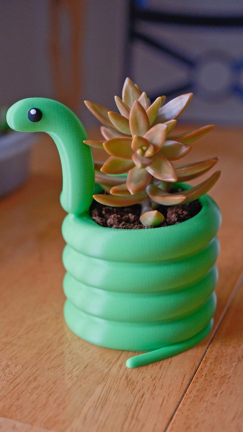 May include: A green snake-shaped planter with a succulent plant. The planter is a vibrant green, coiled to resemble a snake's body, with a head and a small black eye. The succulent has orange and green leaves.