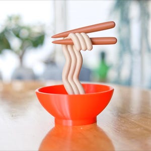 May include: An orange bowl filled with stylized noodles, held by brown chopsticks. The noodles are a light beige color and are arranged in a wavy pattern. The bowl sits on a wooden table, with a blurred background of a window and a plant.