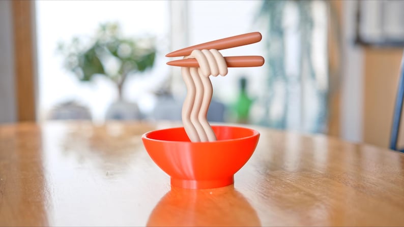 May include: An orange bowl filled with stylized noodles, held by brown chopsticks. The noodles are a light beige color and are arranged in a wavy pattern. The bowl sits on a wooden table, with a blurred background of a window and a plant.