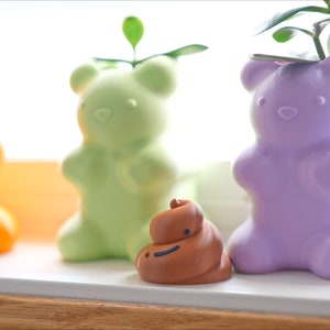May include: Three colorful bear-shaped planters with small plants growing inside. The planters are orange, green, and purple. A small brown figurine of a smiling poop is in front of the planters.