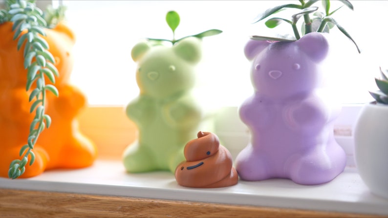 May include: Three colorful bear-shaped planters with small plants growing inside. The planters are orange, green, and purple. A small brown figurine of a smiling poop is in front of the planters.