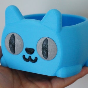 Pode incluir: Um vaso de planta azul claro em forma de gato com olhos cinzentos e um nariz e boca pretos. O vaso &eacute; feito de pl&aacute;stico e tem uma superf&iacute;cie lisa.