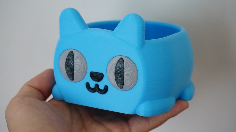 Pode incluir: Um vaso de planta azul claro em forma de gato com olhos cinzentos e um nariz e boca pretos. O vaso &eacute; feito de pl&aacute;stico e tem uma superf&iacute;cie lisa.