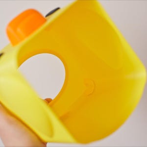 Pu&ograve; includere: Un giocattolo di plastica giallo a forma di anatra con un'ampia apertura al centro. Il giocattolo ha occhi neri e un becco arancione.