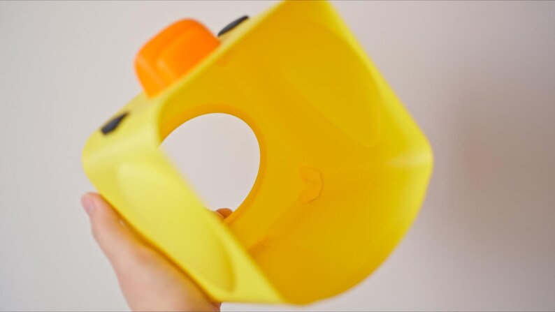Pu&ograve; includere: Un giocattolo di plastica giallo a forma di anatra con un'ampia apertura al centro. Il giocattolo ha occhi neri e un becco arancione.